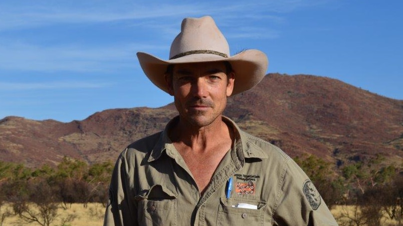 A man in a wide brim hat stands in an outback setting.  He's looking at the camera.