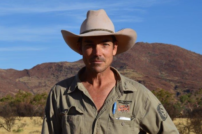 A man in a wide brim hat stands in an outback setting.  He's looking at the camera.