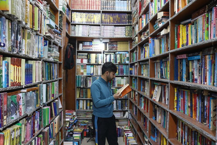 A man is reading a book at a bookshop where bookshelves rise above head height.