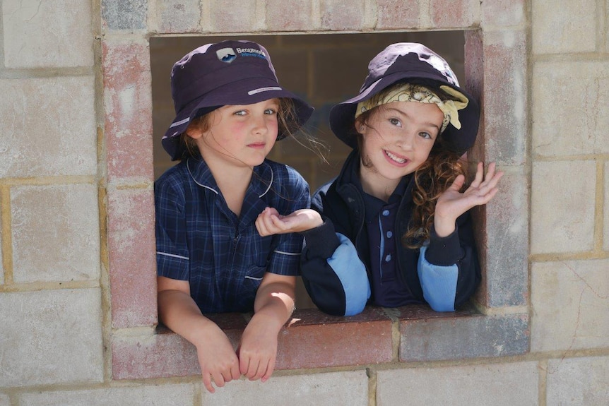 Two girls pose for a photo peeking through an opening in a brick wall in the school yard.