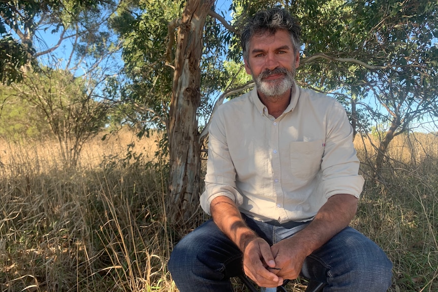 A man in a white buttoned shirt sits in grassy bushland.