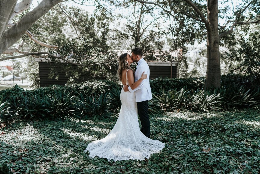 Michelle in a stunning white wedding dress kissing Jono in black pants and white button up shirt, greenery behind.