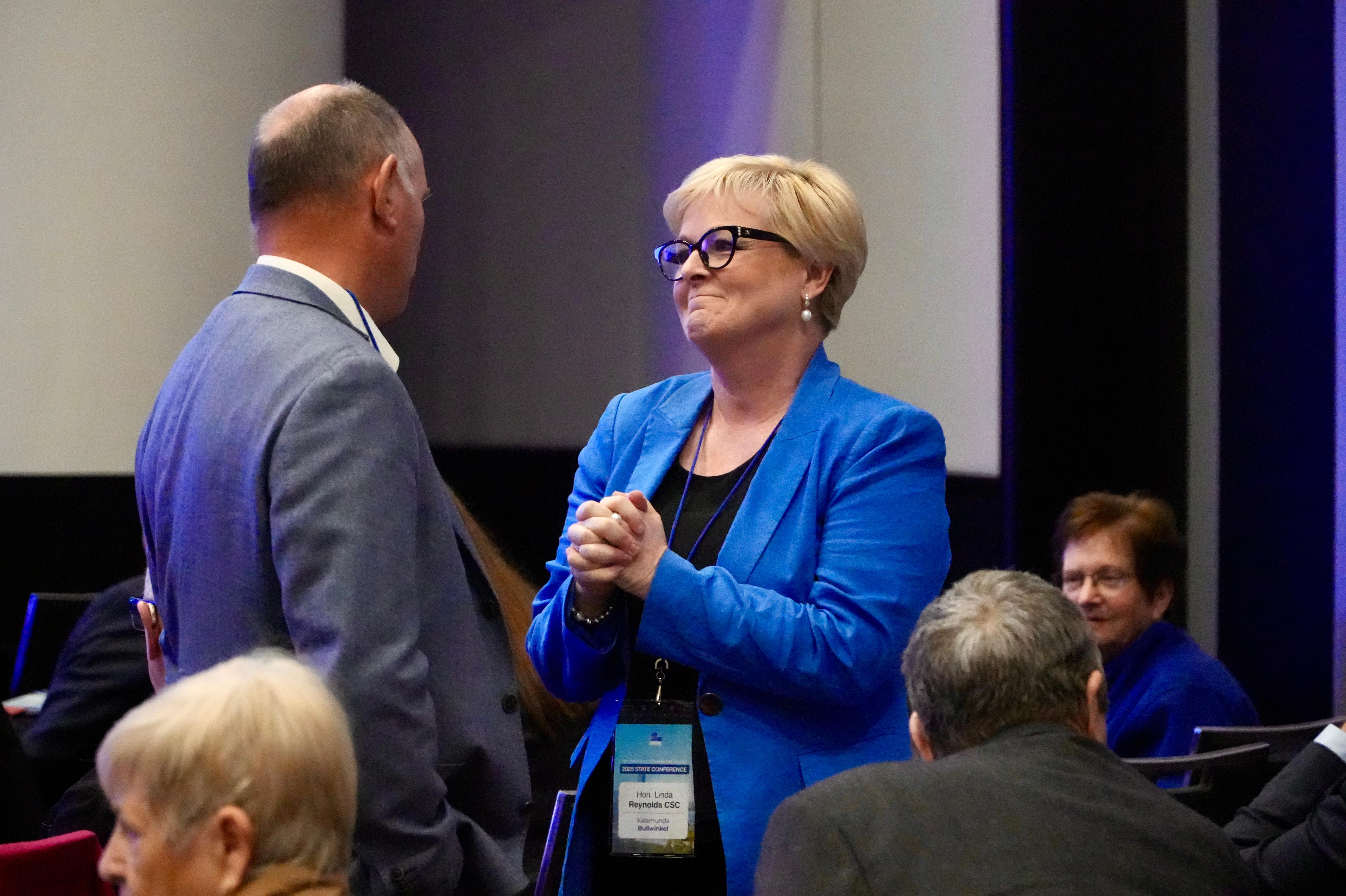 Linda smiles with her hands clasped as she speaks to a man in a grey suit