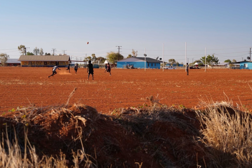 A group of boys play AFL on a red dirt oval