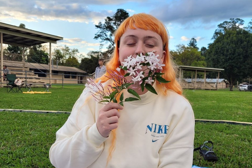A young person with long orange hair smelling a flower in a park