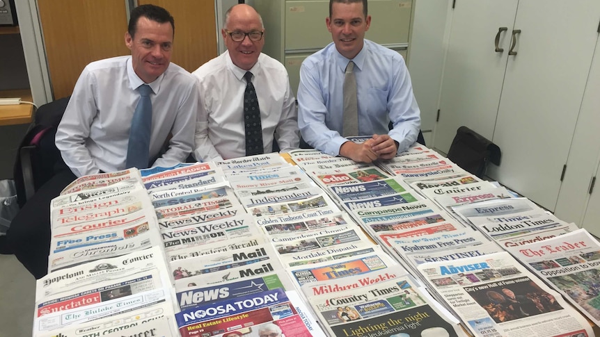 Three men in shirts and ties sitting at a table covered in newspapers