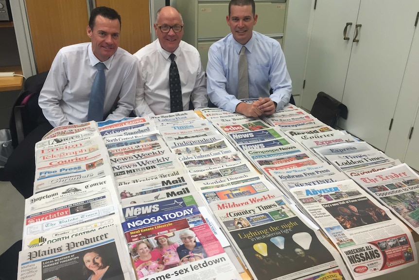 Three men in shirts and ties sitting at a table covered in newspapers