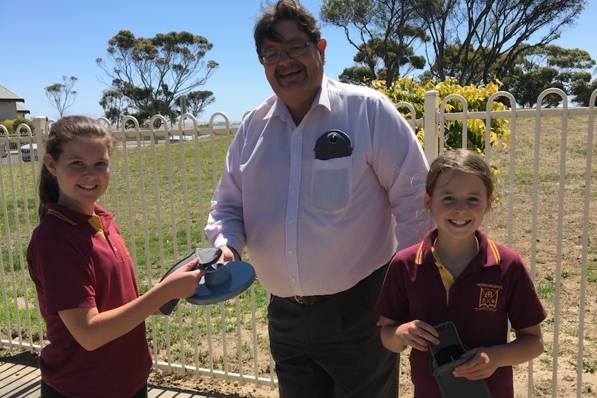 Two female primary school students stand with their principal and test the new mobile phone pouches.