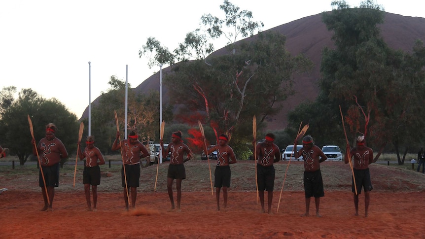 Eight dancers in traditional dress in front of Uluru.