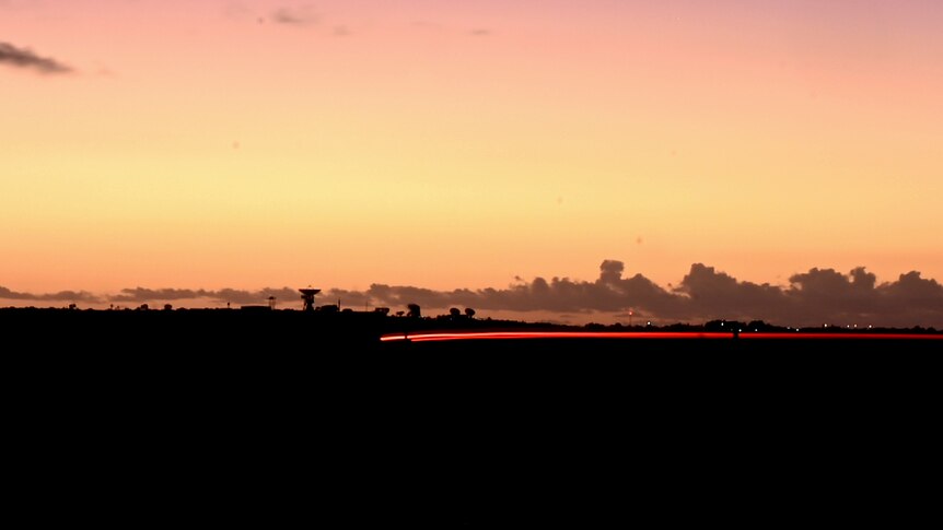 A satellite is silhouetted on a hill and a long orange streak of brake lights is illuminated at sunset.