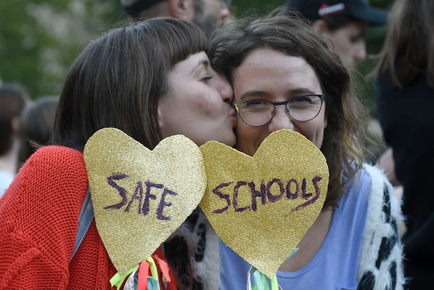 A white person kisses another on the cheek. They are both holding gold, love heart shaped signs that say safe schools.