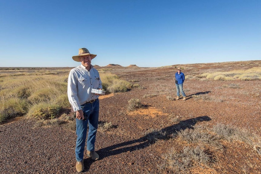 A man and a woman wearing broad-brimmed hats stand in a broad, arid plain with mesas on the horizon.