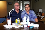 A man and a woman sit at a table looking at the camera. A number of medications sit on the table in front of them.
