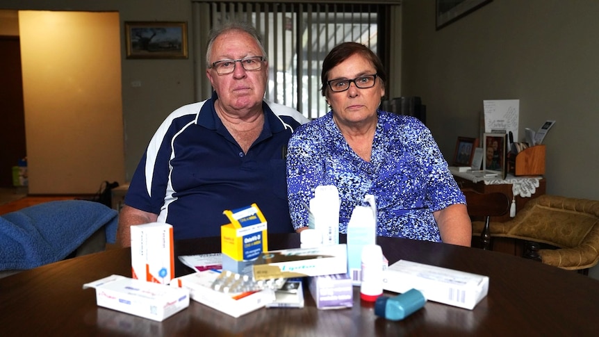 A man and a woman sit at a table looking at the camera. A number of medications sit on the table in front of them.