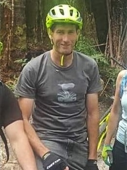 A man wearing a bike helmet, tshirt and jeans sits on his mountain bike and smiles.