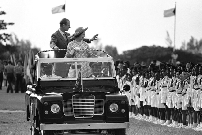 You view an open-top Land Rover Defender carrying the Queen and Prince Philip as they wave to young crowds.