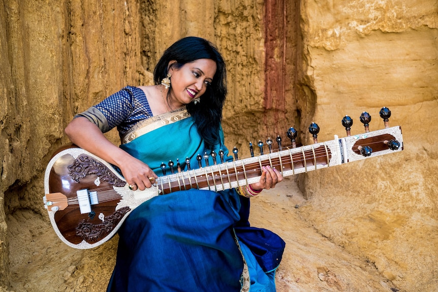 Indian classical sitarist Sarita McHarg sitting in a blue dress and playing her sitar, a plucked string instrument. 