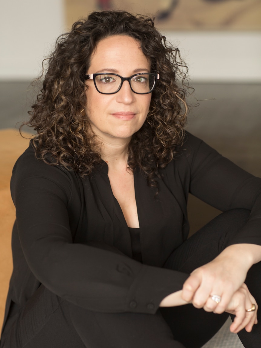 Amy Webb sitting down with hands on knees  wearing reading glasses and a dark outfit, curly hair