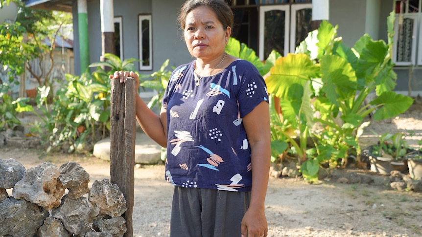 A woman wearing shorts and a top leans on a fence post while standing in her garden.