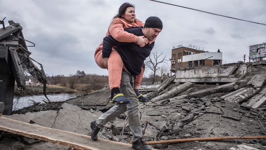 A man carries a woman as they cross a plank above a pile of dark rubble, with a damaged building and canal in the background.