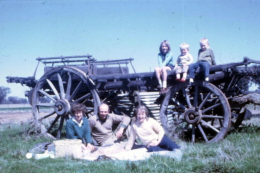 Three adults sitting in front of an old wooden wagon with three children sitting on top of it