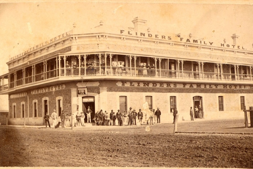 An old black and white photograph of a large, two storey stone building with people gathered by the door and top balcony.