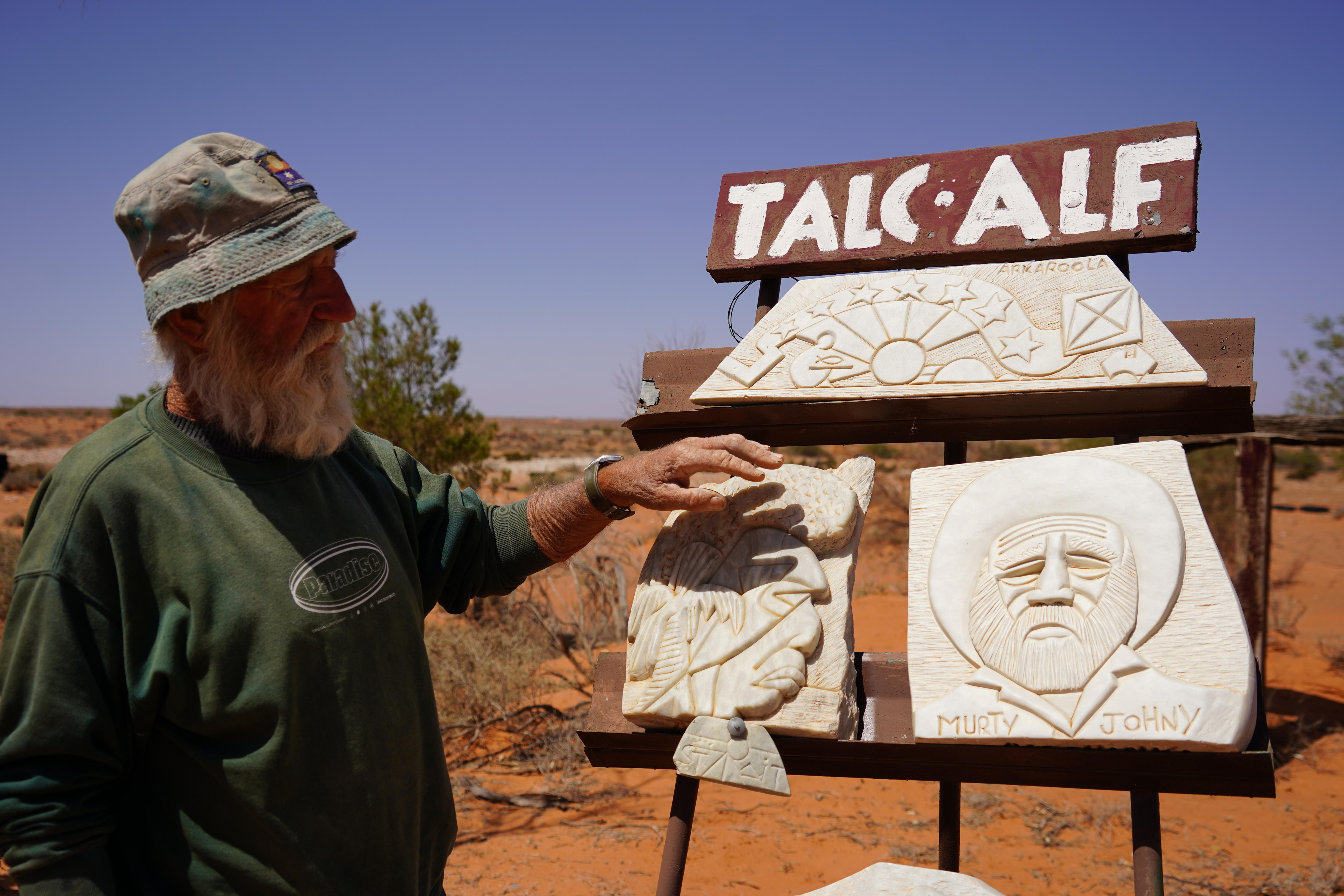 Un hombre con un suéter verde está afuera y mira algunas esculturas de piedra blanca con rostros.