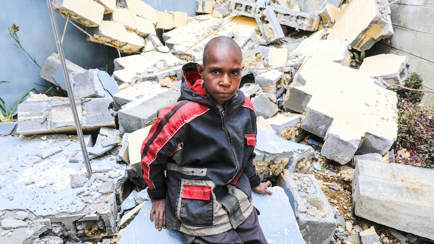 Tight shot of a young boy looking into the camera as he sits on a pile of rubble.