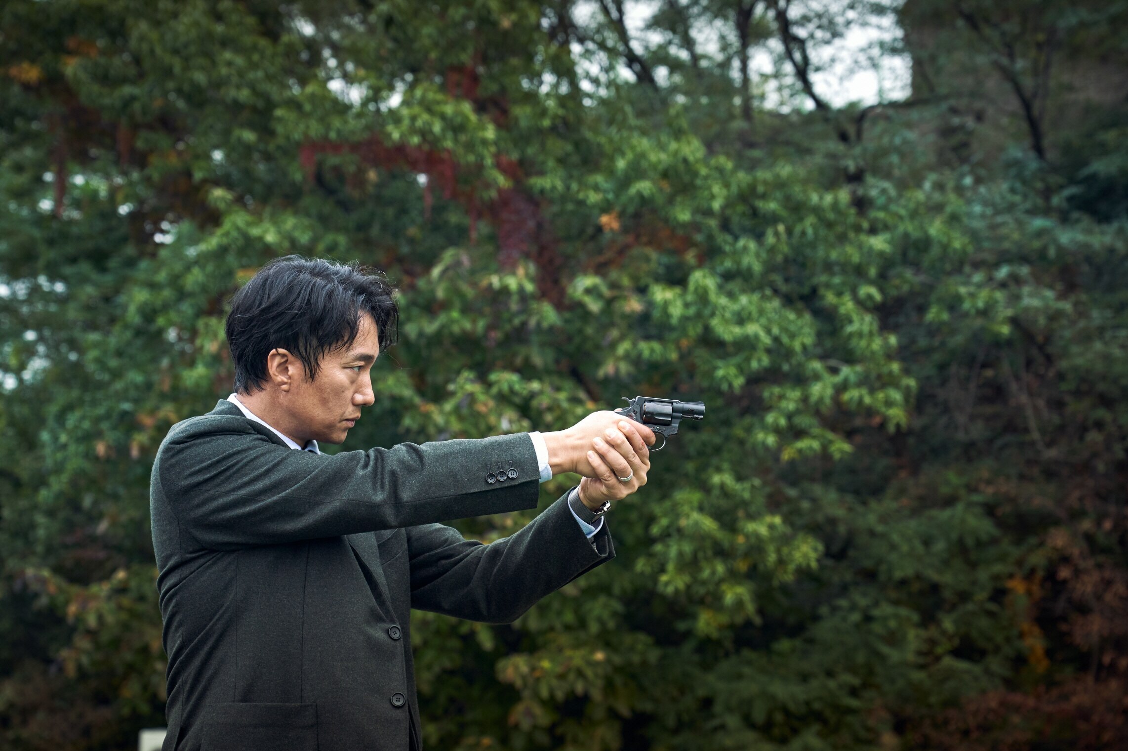 Mid-shot of East Asian man in black suit pointing a piston left to right of frame with lush trees in background.