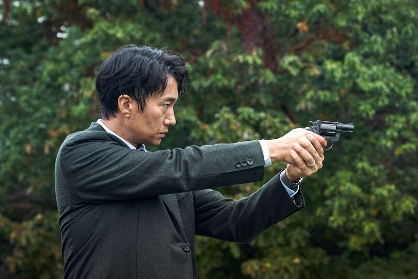 Mid-shot of East Asian man in black suit pointing a piston left to right of frame with lush trees in background.
