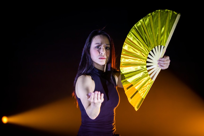 An Asian Australian woman stands on stage, holding a yellow fan up, and with her other hand raised in a beckoning gesture