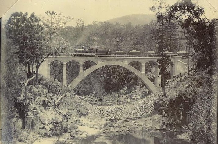 A black and white photo of a locomotive travelling over a concrete bridge above a rocky creek.