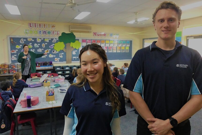A smiling woman and a man in a blue uniform stand in a school classroom as a teacher teaches students.
