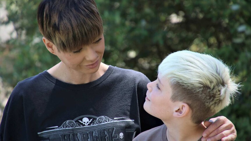 A mid shot of a woman with short hair and a black t-shirt looking down at her young son outdoors.