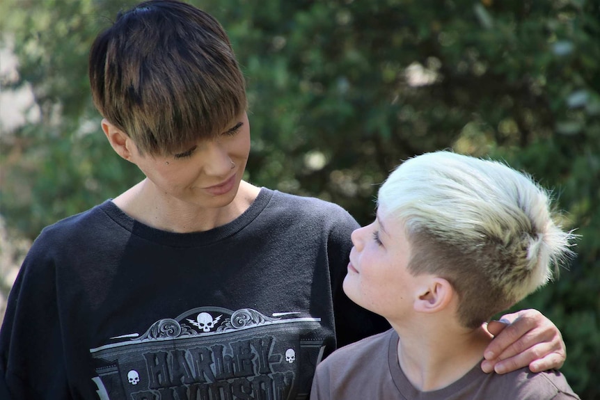 A mid shot of a woman with short hair and a black t-shirt looking down at her young son outdoors.