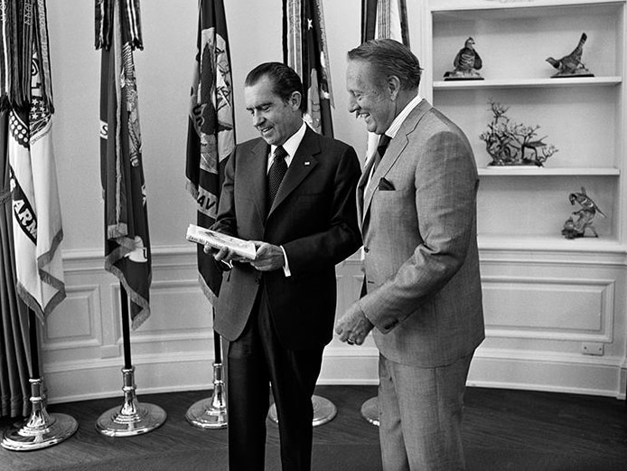 Richard Nixon holds a book in his hands, standing in the Oval Office, while Art Linkletter watches on