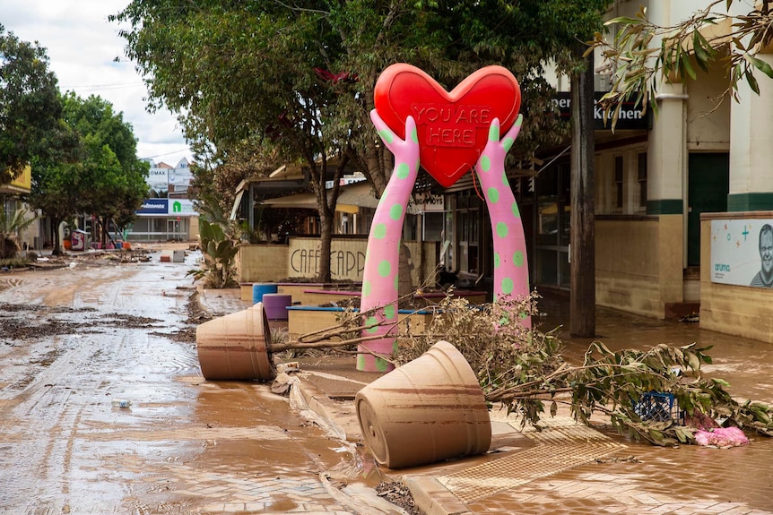 A large public sculpture of a red heart held by two pink and green polka-dotted hands rises above a muddy, flood-ravaged street.