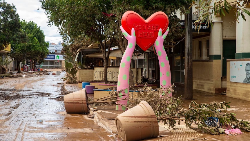 A large public sculpture of a red heart held by two pink and green polka-dotted hands rises above a muddy, flood-ravaged street.