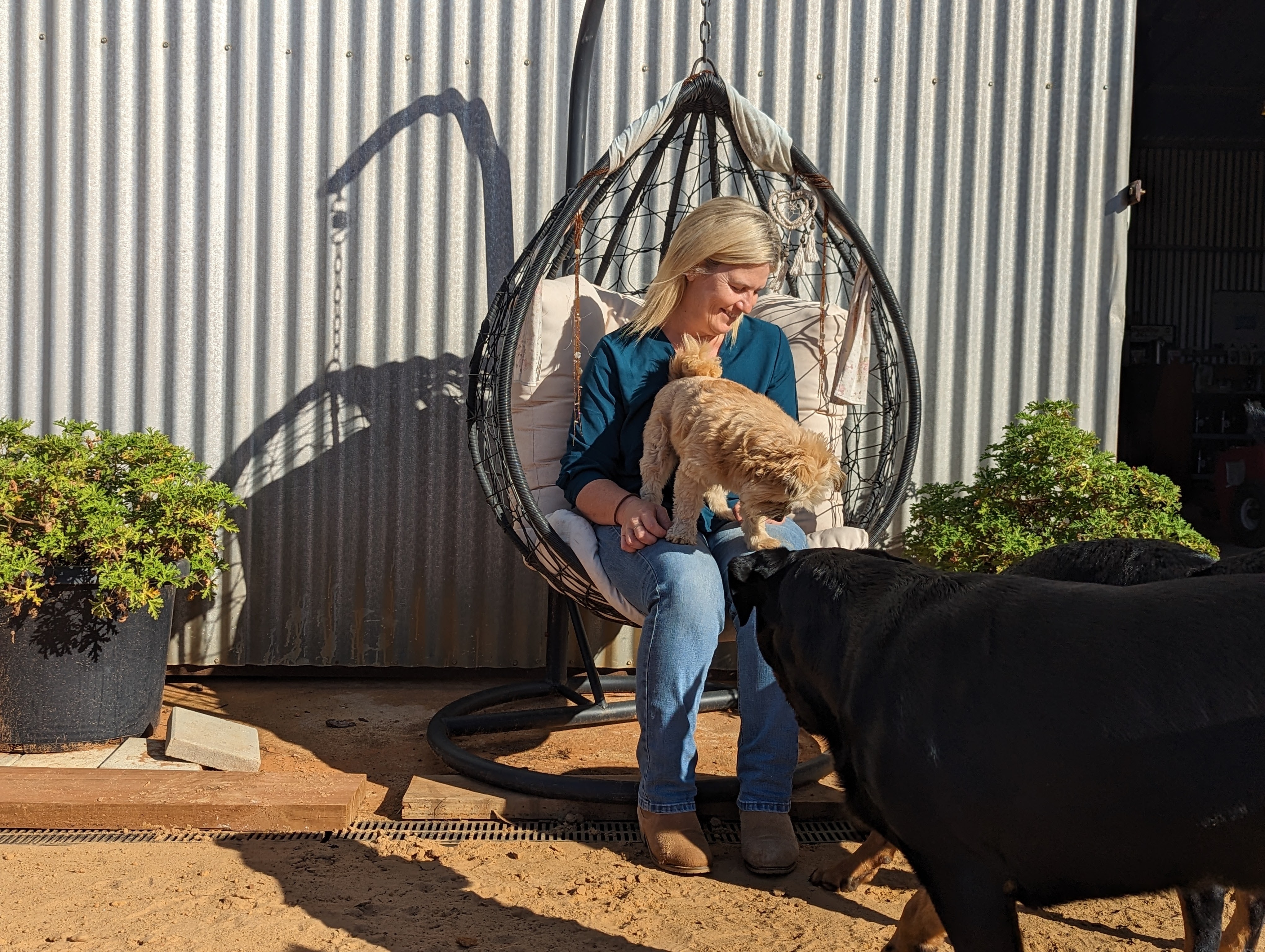 A blonde-haired white woman, Melissa, smiles in an egg chair outside her home with a small fluffy cream dog on her lap. 