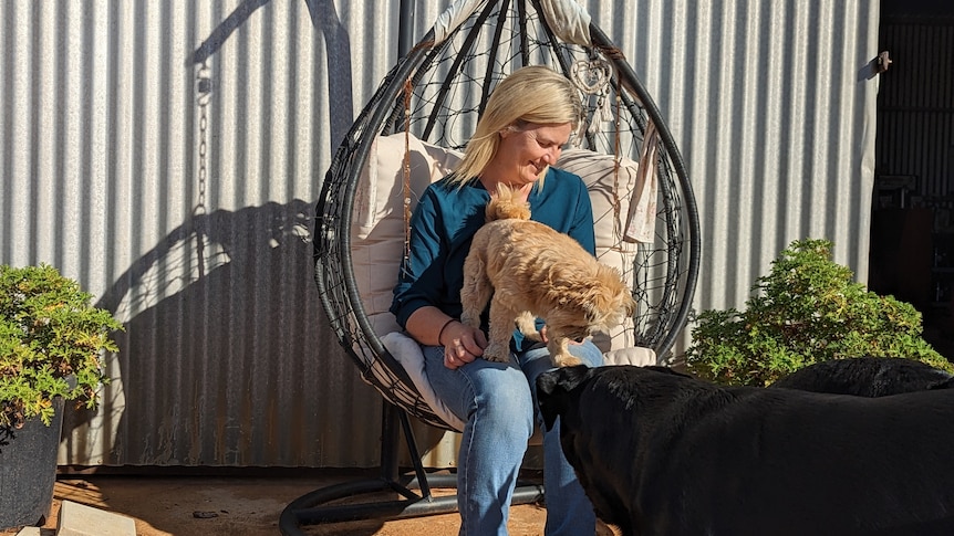A blonde-haired white woman, Melissa, smiles in an egg chair outside her home with a small fluffy cream dog on her lap. 