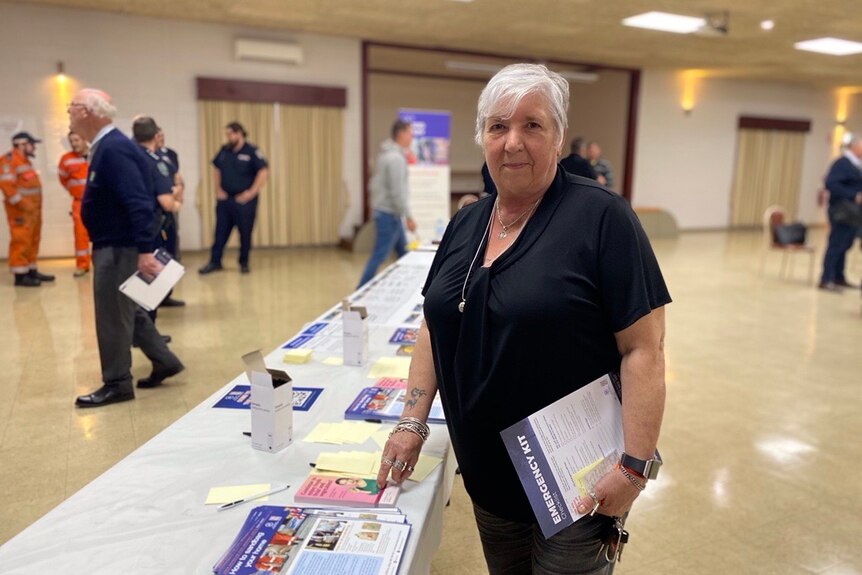 A woman with a black shirt and white hair holds information pamphlets inside a community hall.