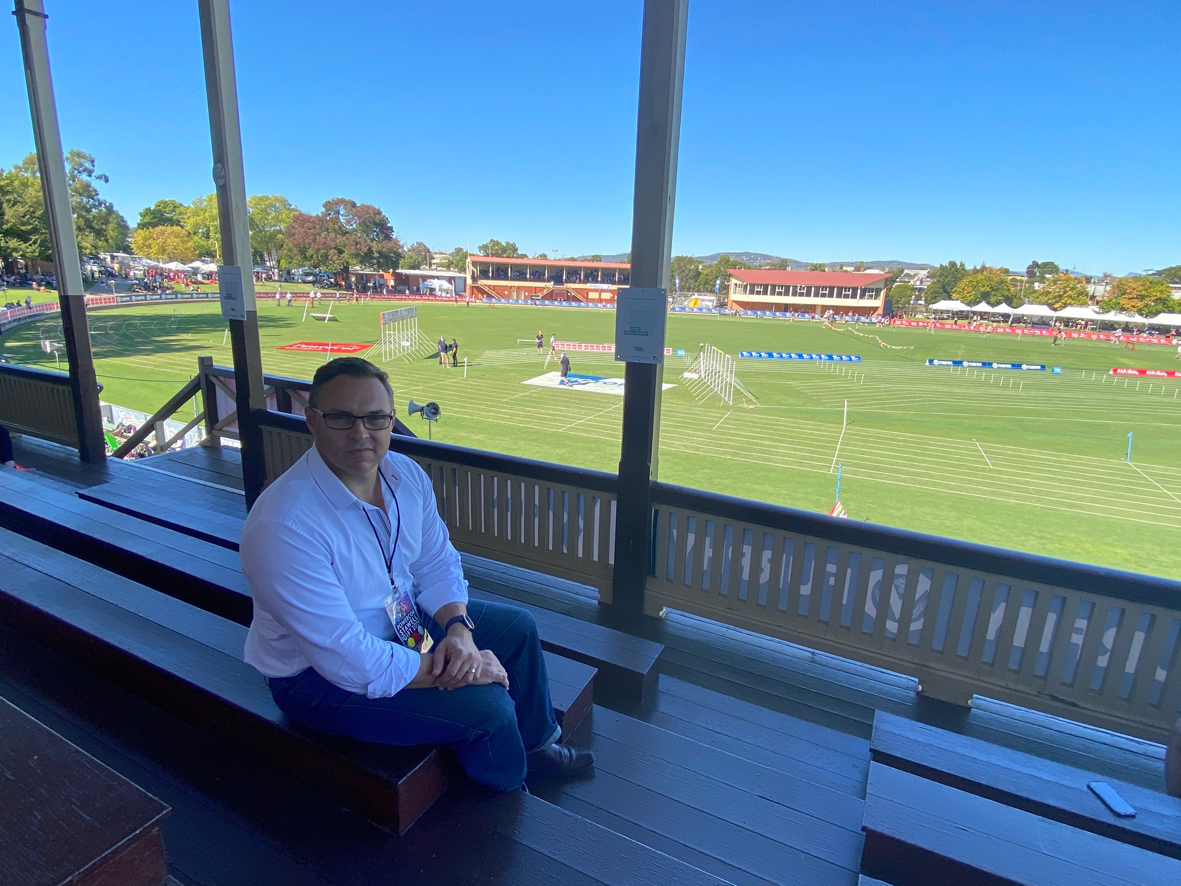 A man in a grandstand looks towards a camera over his shoulder. Behind him is a sunlit athletics track.