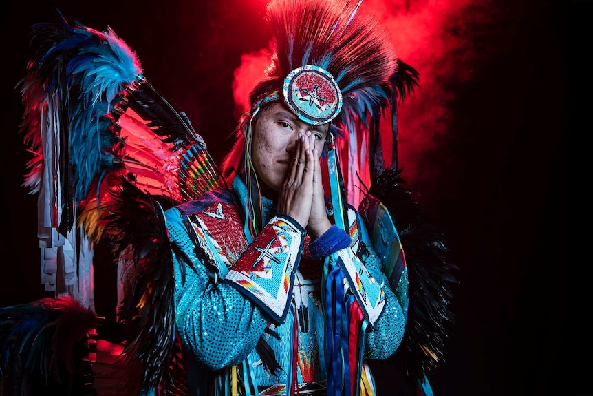 With red smoke behind him, a man in vibrant black, blue and white Native American regalia holds hands in praying pose near face.