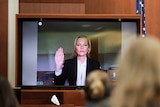 A white woman with blonde hair appears on a monitor raising her right hand with a neutral facial expression