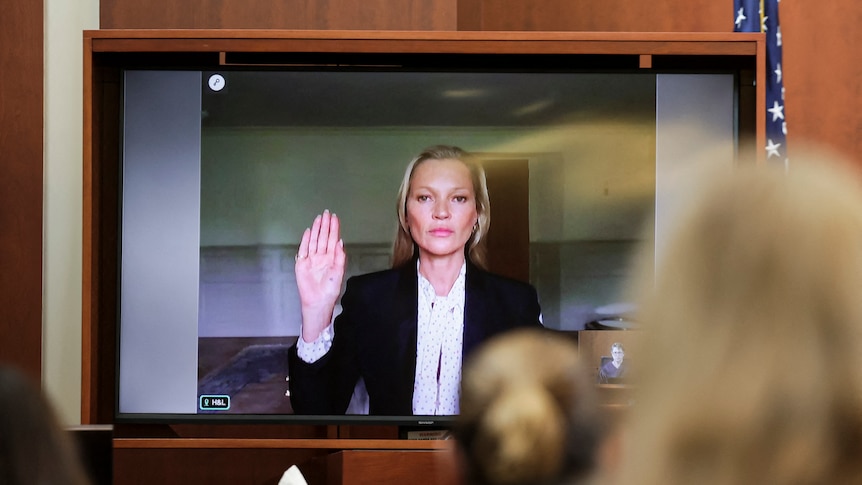 A white woman with blonde hair appears on a monitor raising her right hand with a neutral facial expression