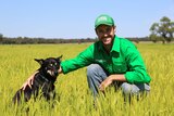 A Kelpie dog sits next to a man wearing a trucker cap and long shirt in a field.