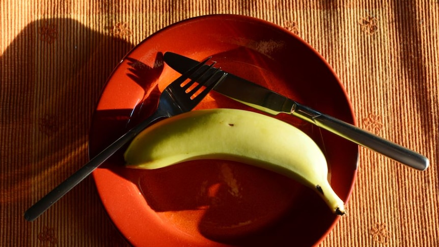 Banana on a red plate with a knife and fork resting on the plate near it.