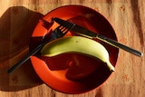Banana on a red plate with a knife and fork resting on the plate near it.