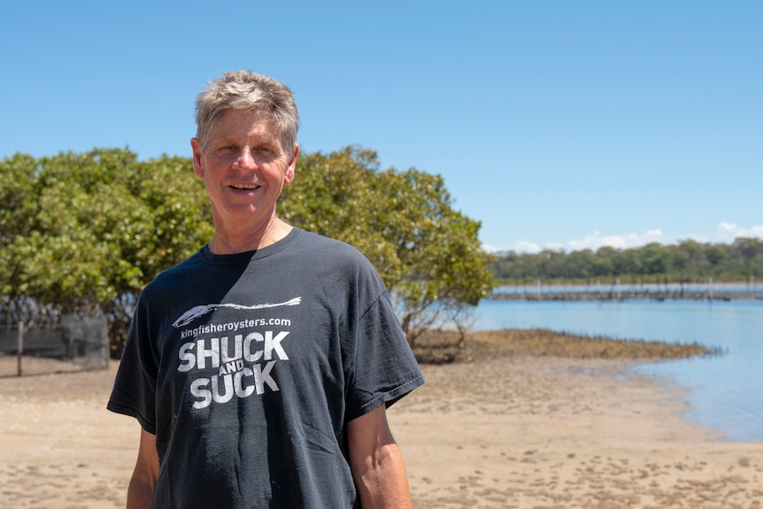 A grey-haired man in a dark t-shirt stands on a sandy riverbank.