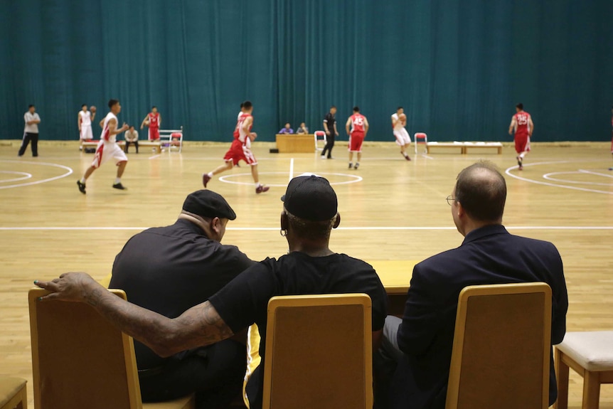 Three men including Dennis Rodman sit and watch a basketball game in North Korea.